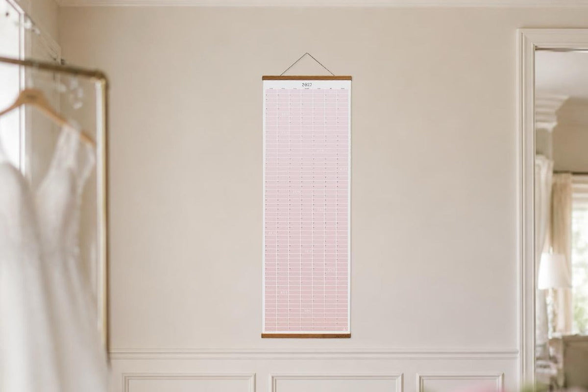 Dahlia Pink 2027 paper wall calendar hanging in a bridal suite beside a wedding dress.