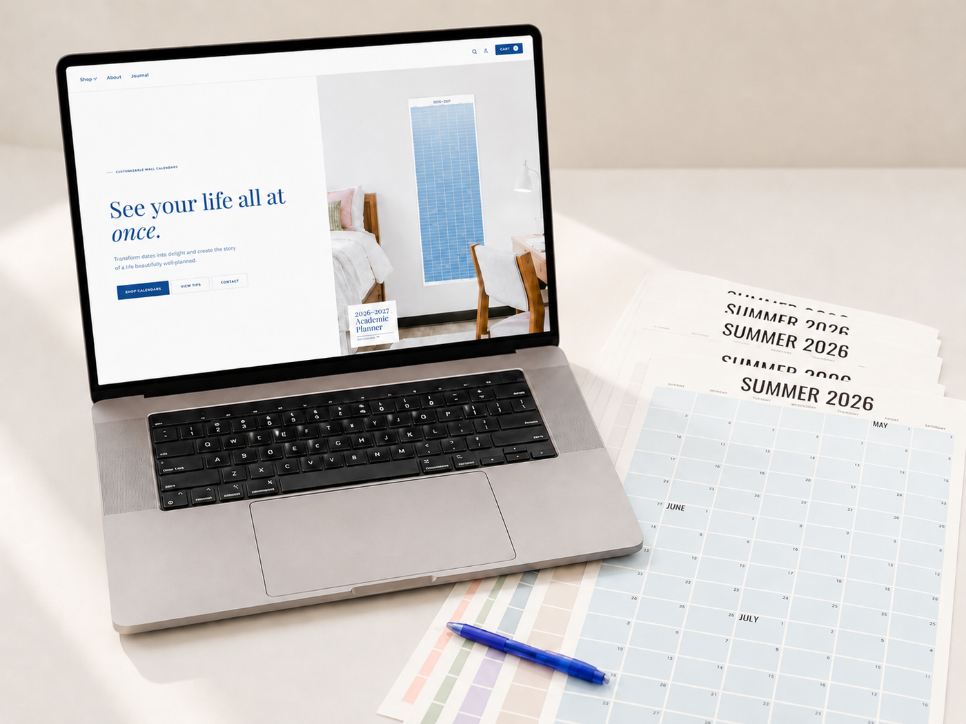 A styled desktop photo showing a laptop open to the Pecan Print House website, with printed Summer 2026 calendar pages and a blue pen arranged on a clean, neutral studio surface.