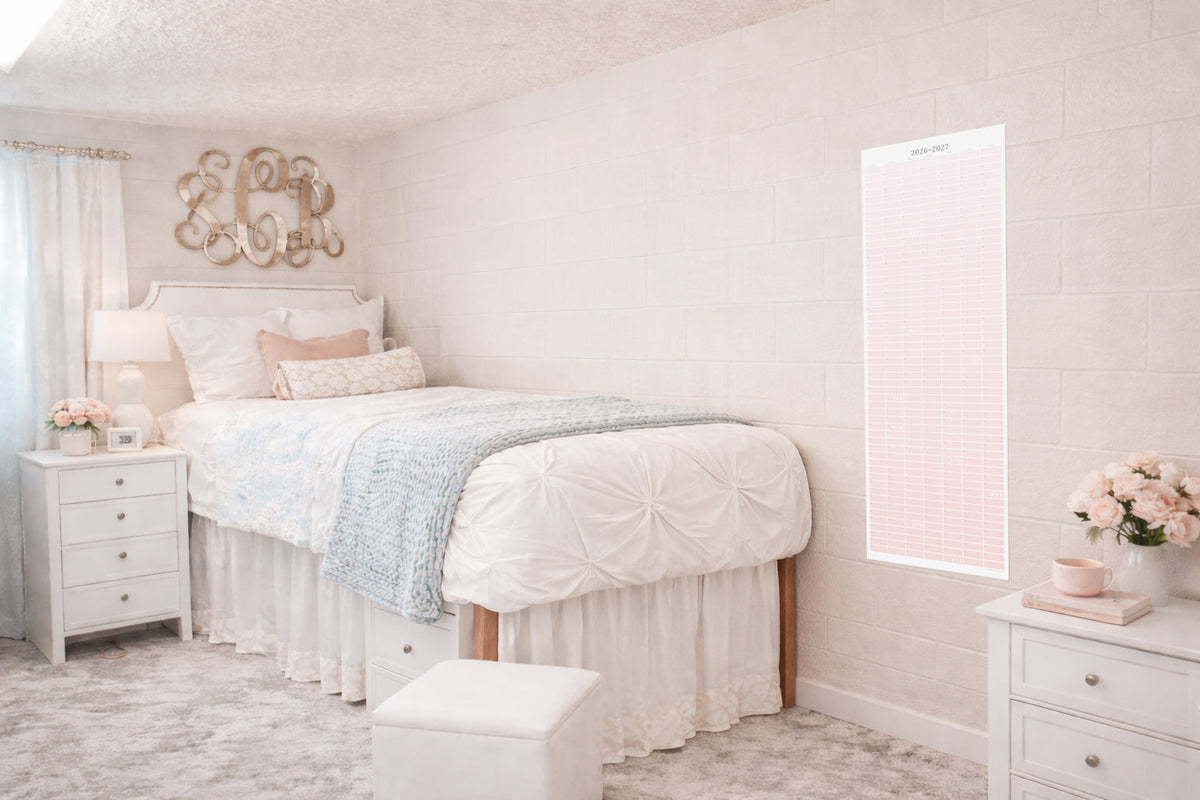 Soft blush dorm bedroom with a pink 2026–2027 academic wall calendar hanging on a light brick wall beside a neatly styled bed, white bedding, and pastel decor, showcasing a decorative continuous school year planner for college students.