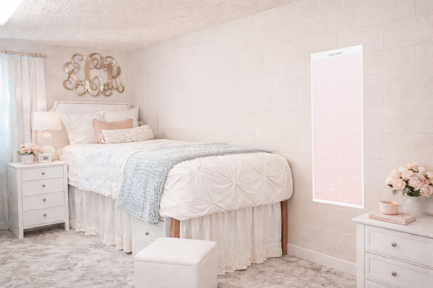 Soft blush dorm bedroom with a pink 2026–2027 academic wall calendar hanging on a light brick wall beside a neatly styled bed, white bedding, and pastel decor, showcasing a decorative continuous school year planner for college students.