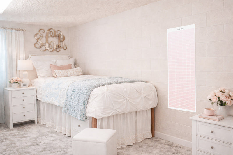 Soft blush dorm bedroom with a pink 2026–2027 academic wall calendar hanging on a light brick wall beside a neatly styled bed, white bedding, and pastel decor, showcasing a decorative continuous school year planner for college students.
