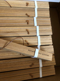 A set of natural wood sticks bundled with white elastic bands and tied with small leather loops, displayed inside a cardboard box. One stick is leaning diagonally and features the printed brand name 'PECAN PRINT HOUSE'.