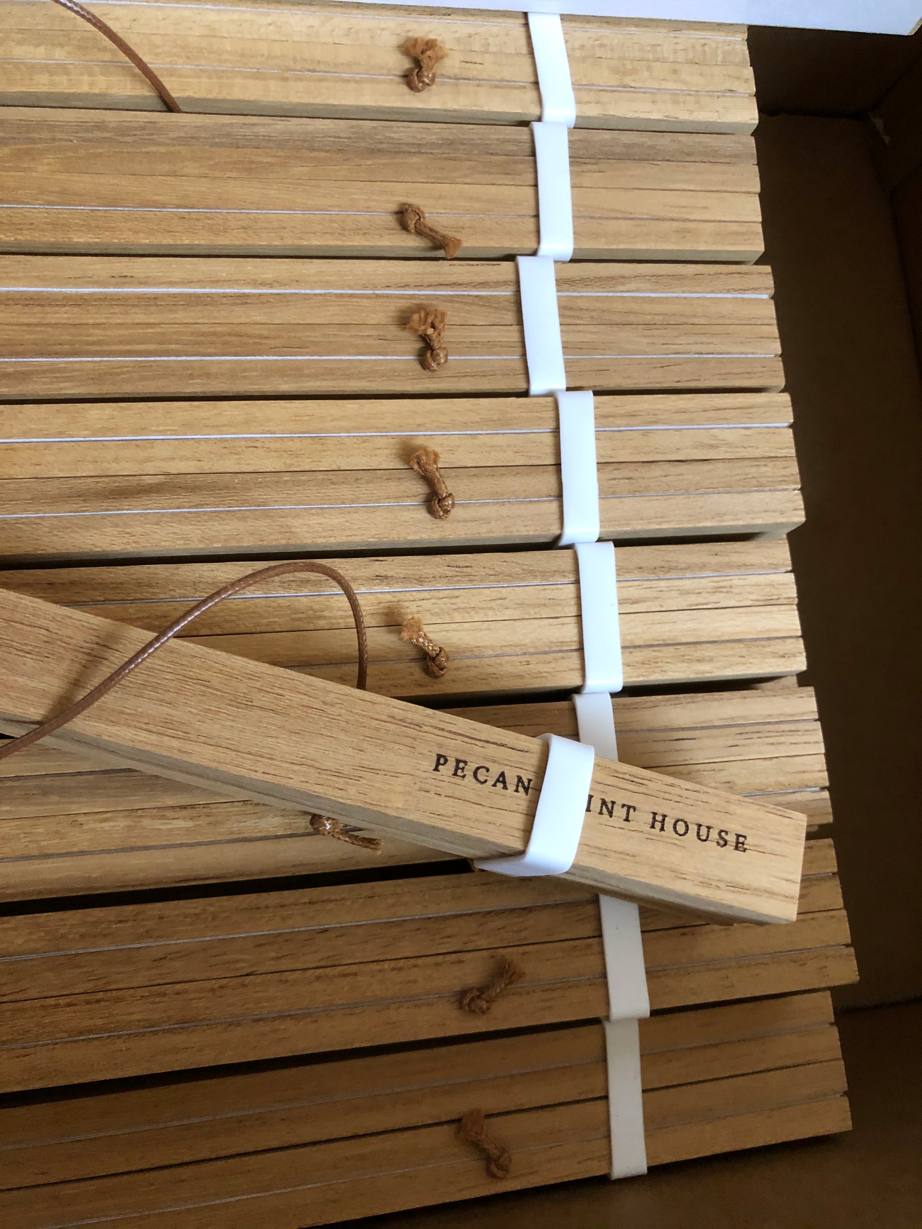 A set of natural wood sticks bundled with white elastic bands and tied with small leather loops, displayed inside a cardboard box. One stick is leaning diagonally and features the printed brand name 'PECAN PRINT HOUSE'.