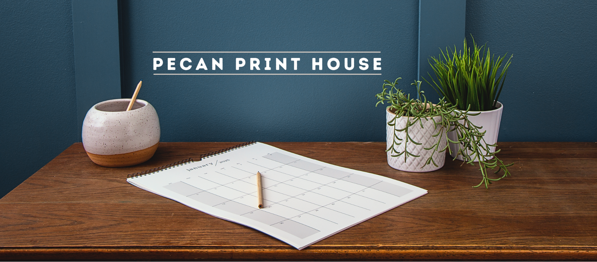 Desk setup with notebook, pen, and plants against a blue wall with 'Pecan Print House' branding.
